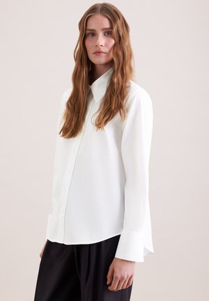 Kvinde med langt bølget hår iført en hvid skjorte med knapper og sorte bukser, stående mod en enkel lys baggrund.