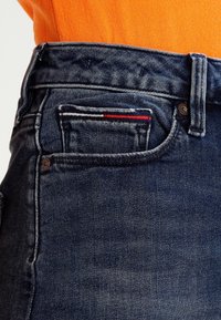 Tmavě modré džíny z denimu s klasickým designem, který zahrnuje malou přední kapsu lemovanou červenými a bílými pruhy. Na povrchu je vybledlá textura.