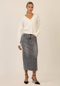 Cardigan bianco a coste con scollo a V e bottoni frontali, abbinato a una gonna lunga in denim grigia con dettagli in strass e tacchi neri.