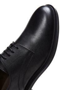 Zwarte leren schoen met een gladde textuur, vetersluiting, ronde neus en gestepte accenten langs de zijkanten en de zool.