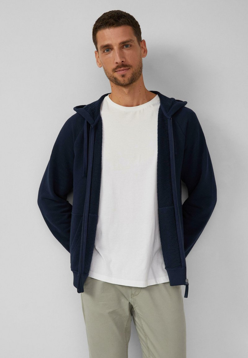 Felpa blu navy con zip frontale, tessuto testurizzato, coulisse e due tasche frontali, indossata sopra una t-shirt bianca e pantaloni beige chiaro.