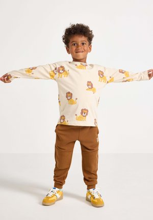 Crème lange mouwen shirt met leeuwendruk, gecombineerd met bruine joggers met ribbelaccenten en gele sneakers. Glad materiaal, ontspannen pasvorm.