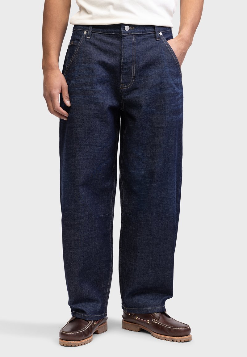 Jeans din denim de culoare albastru închis, cu o croială relaxată, dotate cu buzunare frontale, cusături vizibile și închidere cu nasture. Purtați cu pantofi din piele maronie.