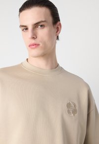 Beige pulóver bordázott textúrával és kerek nyakkivágással, finoman hímzett logóval a bal mellen. Simogató, könnyű anyag.