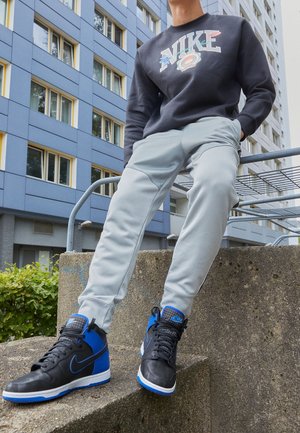 Osoba nosząca czarne i niebieskie wysokie buty sportowe Nike, jasnoszare spodnie i czarną bluzę Nike, siedząca na zewnętrznej betonowej konstrukcji.