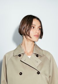 Cappotto beige in stile trench, con design doppiopetto, bottoni scuri e collo a reverse; tessuto liscio con caratteristiche minimaliste e cuciture sottili.