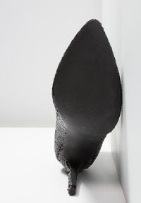 Chaussure à talon haut noire à bout pointu ornée de sequins, avec une semelle texturée, photographiée par en dessous sur un fond blanc.
