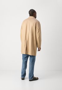 Beige lange jas met een relaxte pasvorm, rugopening en puntkraag, gecombineerd met blauwe jeans en zwart schoeisel. Gladde textuur en minimalistisch ontwerp.