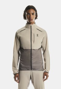 Jeune personne avec des cheveux bouclés de longueur moyenne portant une veste zippée beige et grise ainsi qu'un pantalon assorti, se tenant devant un fond blanc.