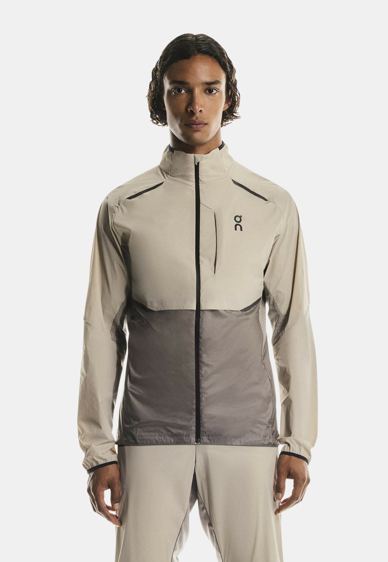 Jeune personne avec des cheveux bouclés de longueur moyenne portant une veste zippée beige et grise ainsi qu'un pantalon assorti, se tenant devant un fond blanc.