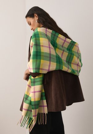 Žena s dlhými tmavými vlasmi zabalená v šále so zeleným, žltým a fialovým károvaným vzorom cez hnedý kabát, pozerajúca sa dole na neutrálnom pozadí.