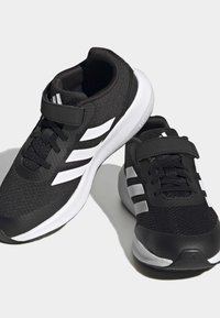 Zapatillas deportivas negras con parte superior de tejido texturizado, acentos blancos y una correa de gancho y bucle. Cuenta con una suela acolchada y rayas en contraste.