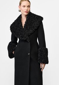 Czarny wełniany płaszcz z dużym, teksturowanym futrzanym kołnierzem i dopasowanymi futrzanymi akcentami na rękawach. Zapięcie na jeden guzik, dopasowany krój.