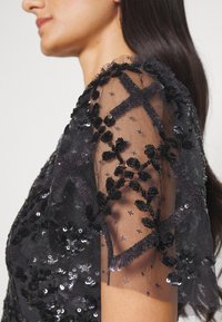 Femme portant un haut en dentelle noire transparente avec une broderie de sequins florale et géométrique sur la manche, montrant le profil de côté et de longs cheveux noirs.