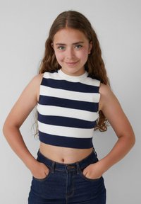 Gestreiftes ärmelloses Crop-Top in Marineblau und Weiß aus geripptem Strickstoff, kombiniert mit hoch taillierten Jeans aus Denim, Hände in den Taschen.