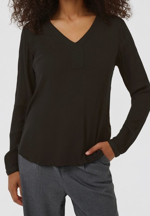 Femme portant un chemisier noir à manches longues et col en V avec un pantalon gris, une main dans la poche, sur un fond uni.