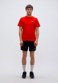 Κόκκινο βαμβακερό μπλουζάκι με κοντά μανίκια, μικρό λευκό λογότυπο· συνδυασμένο με μαύρο σορτς, λευκές κάλτσες και μαύρα αθλητικά παπούτσια.