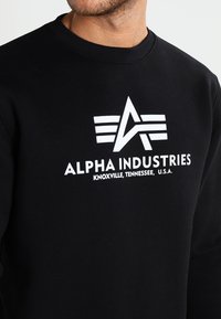 Sudadera negra con cuello redondo que presenta un gran logotipo blanco que incluye letras estilizadas y rayas horizontales.