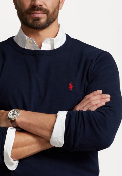 Suéter azul marino con cuello redondo, tejido texturizado y un pequeño logotipo rojo bordado. Las mangas del suéter muestran puños de camisa blanca debajo.