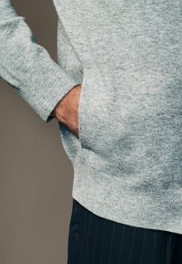 Jasnoszary sweter o fakturowanym wykończeniu, z boczną kieszenią i ściągaczami na rękawach. Połączony z ciemnymi spodniami w prążki.