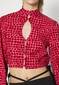 Blusa curta vermelha com gola alta, abertura em buraco de chave e fecho com botão. O tecido apresenta um padrão texturizado com formas circulares variadas.