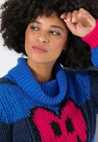 Strickpullover mit blauem Schalkragen, auffälligem Herzdesign in Pink und Schwarz sowie strukturierten Mustern im Stoff.