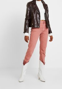 Braune Bikerjacke aus Kunstleder, weißes geripptes Crop-Top, pinke hoch taillierte Jeans und weiße Cowgirl-Stiefel mit strukturiertem Detail.