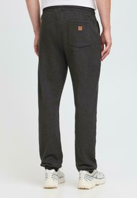 Mann iført mørkegrå joggebukser med baklomme og hvite joggesko, står mot en ensfarget lys bakgrunn.