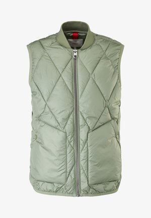 Uomo che indossa un gilet trapuntato verde chiaro, camicia a maniche lunghe a righe color crema, pantaloni color cammello con risvolti e scarpe da ginnastica bianche, in piedi su un pavimento beige.