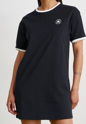 Kvinde iført en sort kortærmet kjole med hvid kant og et lille Converse All Star-logo på brystet.