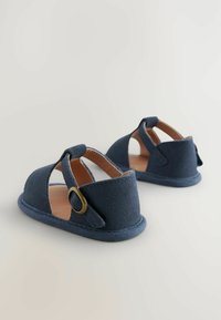 Sandali blu navy realizzati in morbida suede, con lati intagliati, una cintura con fibbia tonda e una suola piatta. Dettagli di design fatti a mano visibili.