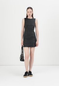 Zwart denim mouwloos jurkje met een getailleerde pasvorm, voornaaddetails en een zijrits. Gecombineerd met zwarte sneakers en een kleine handtas.
