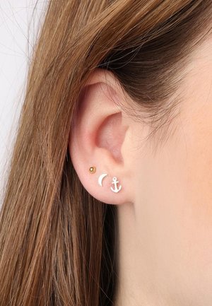 Trois boucles d'oreilles : une boule en or, un croissant de lune blanc et une ancre blanche, posées sur une oreille lisse. Des cheveux bruns cachent partiellement l'oreille.