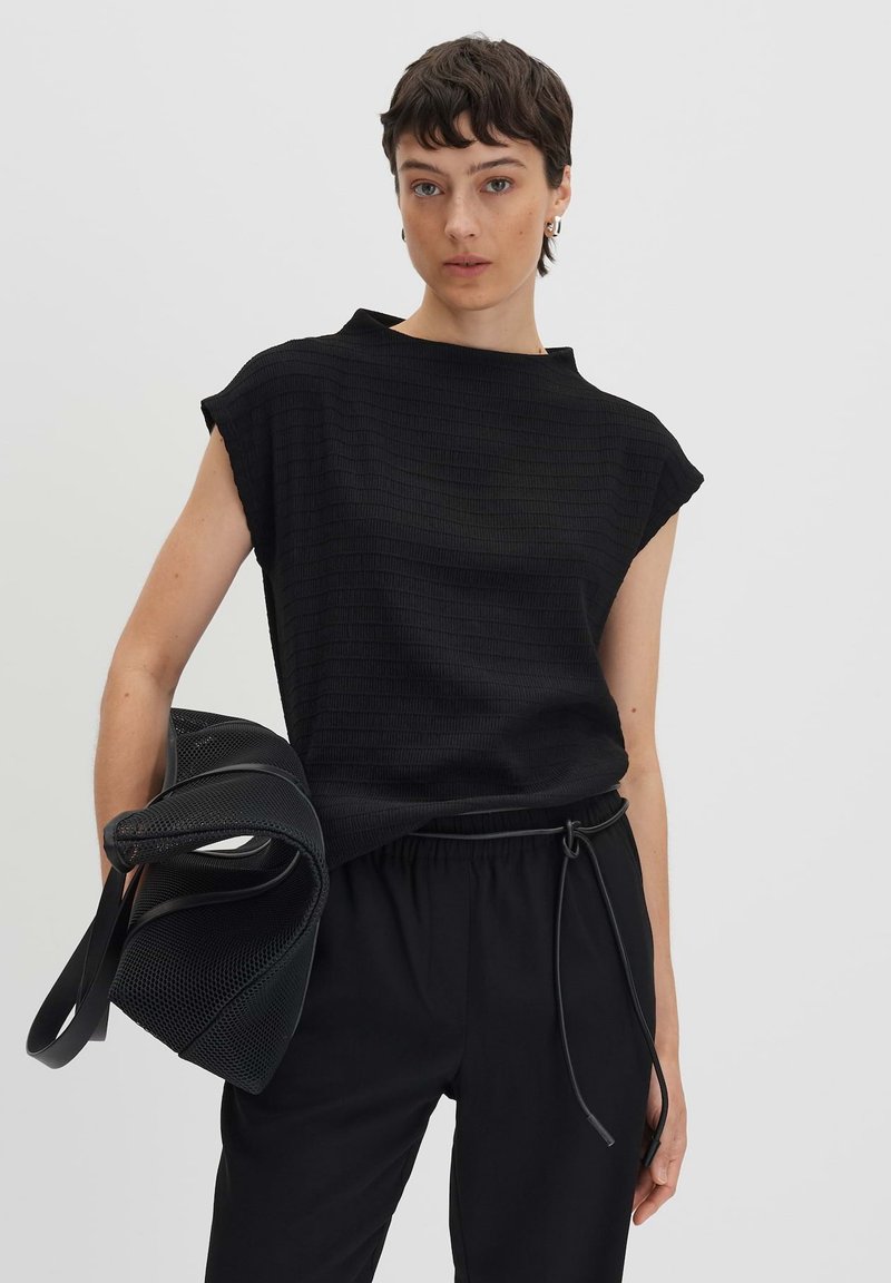Schwarzes, strukturiertes Oberteil mit hohem Kragen und kurzen Ärmeln, kombiniert mit schwarzen Hosen mit elastischem Bund und einer schwarzen Netztragetasche.
