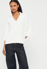Pulôver branco de malha com decote em V e mangas curtas largas, combinado com uns jeans azul escuro. O tecido parece suave e com uma textura lisa.