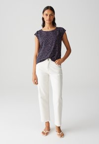 Blusa navy con pois colorati, maniche corte e scollo rotondo; abbinata a jeans bianchi a gamba dritta e infradito beige.