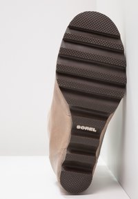 Stivale in suede marrone con suola in gomma nera texturizzata caratterizzata da profonde scanalature e logo "SOREL". Lo stivale ha una finitura liscia e punta arrotondata.