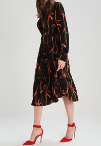 Schwarzes Kleid mit rotem und orangefarbenem Blumenmuster, langen Ärmeln, tailliertem Bund, fließendem Stoff; kombiniert mit roten High Heels mit Knöchelriemen.