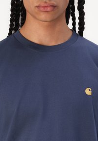 Navy blue cotton t-shirt featuring a crew neck and a small gold logo on the left chest. Smooth texture with short sleeves.