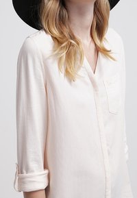 Blusa lunga a maniche con scollo a V di colore rosa chiaro, dotata di una tasca sul petto e maniche arrotolate, realizzata in un tessuto morbido e liscio.