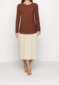 Haut marron à manches longues côtelées associé à une jupe midi plissée beige et des sandales à lanières marron clair, présenté sur un fond blanc uni.