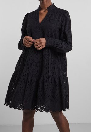 Czarna sukienka wykonana z tkaniny broderie anglaise, z dekoltem w serek, długimi rękawami, tiulowym dołem i falistymi krawędziami.