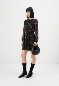 Femme avec une coupe au carré noir portant une robe longue transparente à fleurs noires, des chaussettes noires, des chaussures à talons, et tenant un sac à main noir.