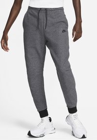 Šedé fleecové tepláky se zúženými nohavicemi, černý pas a manžety, s logem na horní části stehna. Nošeny s bílými sportovními botami.