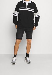 Camisa negra de manga larga con rayas horizontales blancas y grises, que presenta un collar con cremallera. Combinada con pantalones cortos de mezclilla negros remangados y zapatillas grises.