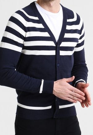 Mężczyzna w granatowym kardiganie z białymi poziomymi paskami na białym t-shircie, dłonie złożone z przodu.