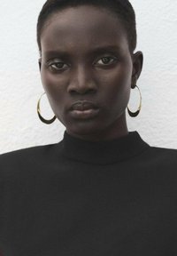 Černý top s vysokým límcem, s hladkým povrchem. Model nosí zlaté kruhové náušnice, které zdobí odvážný a minimalistický design na světlém pozadí.