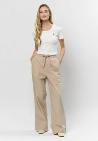 Biely rebrovaný tričko s malým logom, skombinované s béžovými nohavicami s širokými nohavicami, elastickým pásom a zaviazaným detailom, stojace na jednofarebnom pozadí.