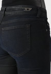 Takana näkymä tummansinisistä farkuista, joissa on näkyvä takatasku ja metallinen logo vyötäröllä vaaleaa taustaa vasten.