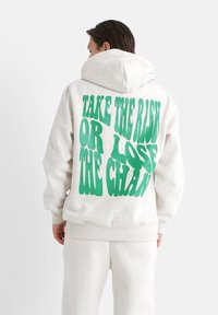 Sudadera gris claro con un gran texto gráfico verde en la parte posterior: "TOMA EL RIESGO O PIERDE LA OPORTUNIDAD." Con capucha ajustable y puños acanalados.